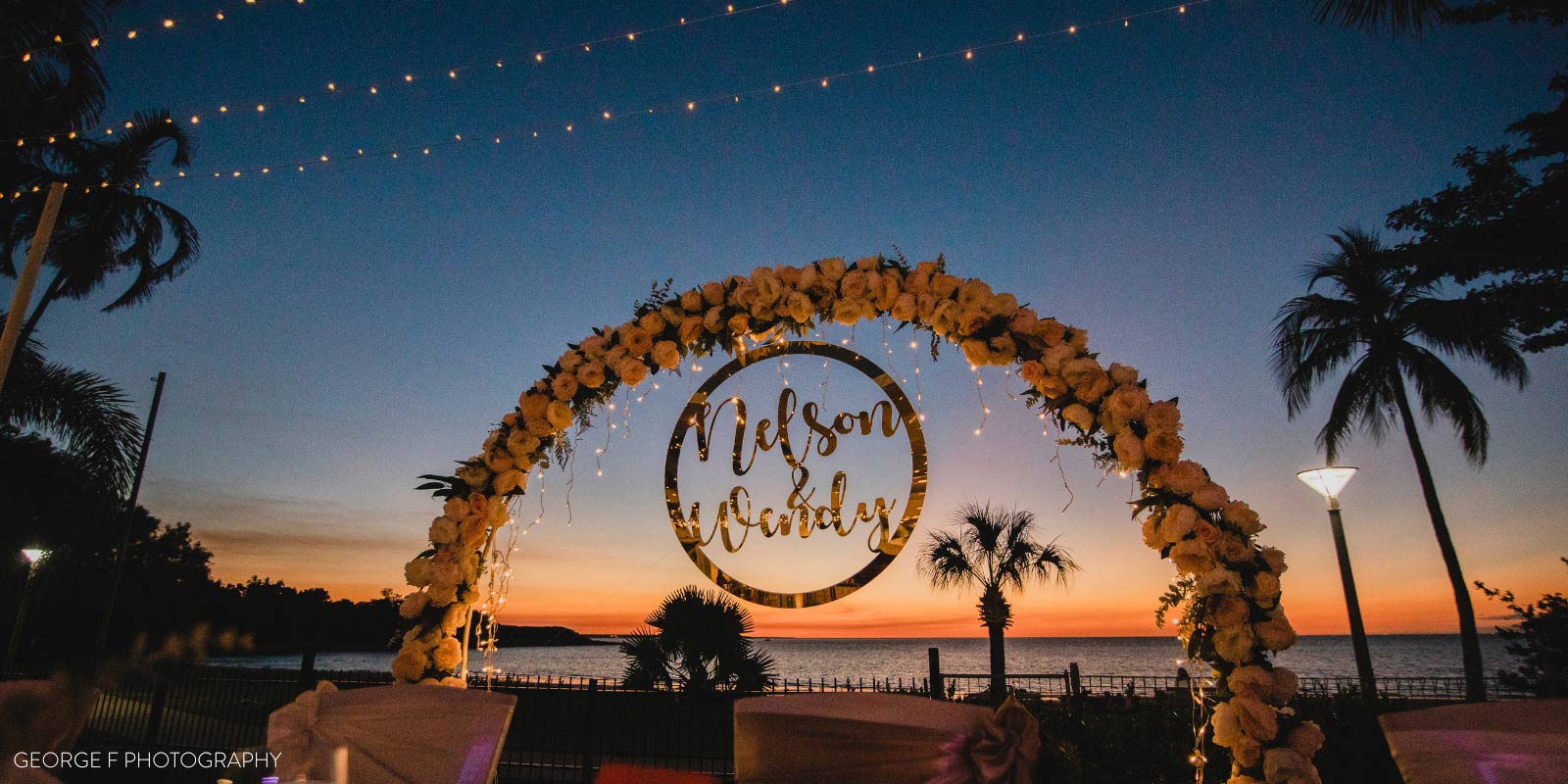 Photo of wedding arch at dusk featuring palm trees and string lights and flowers