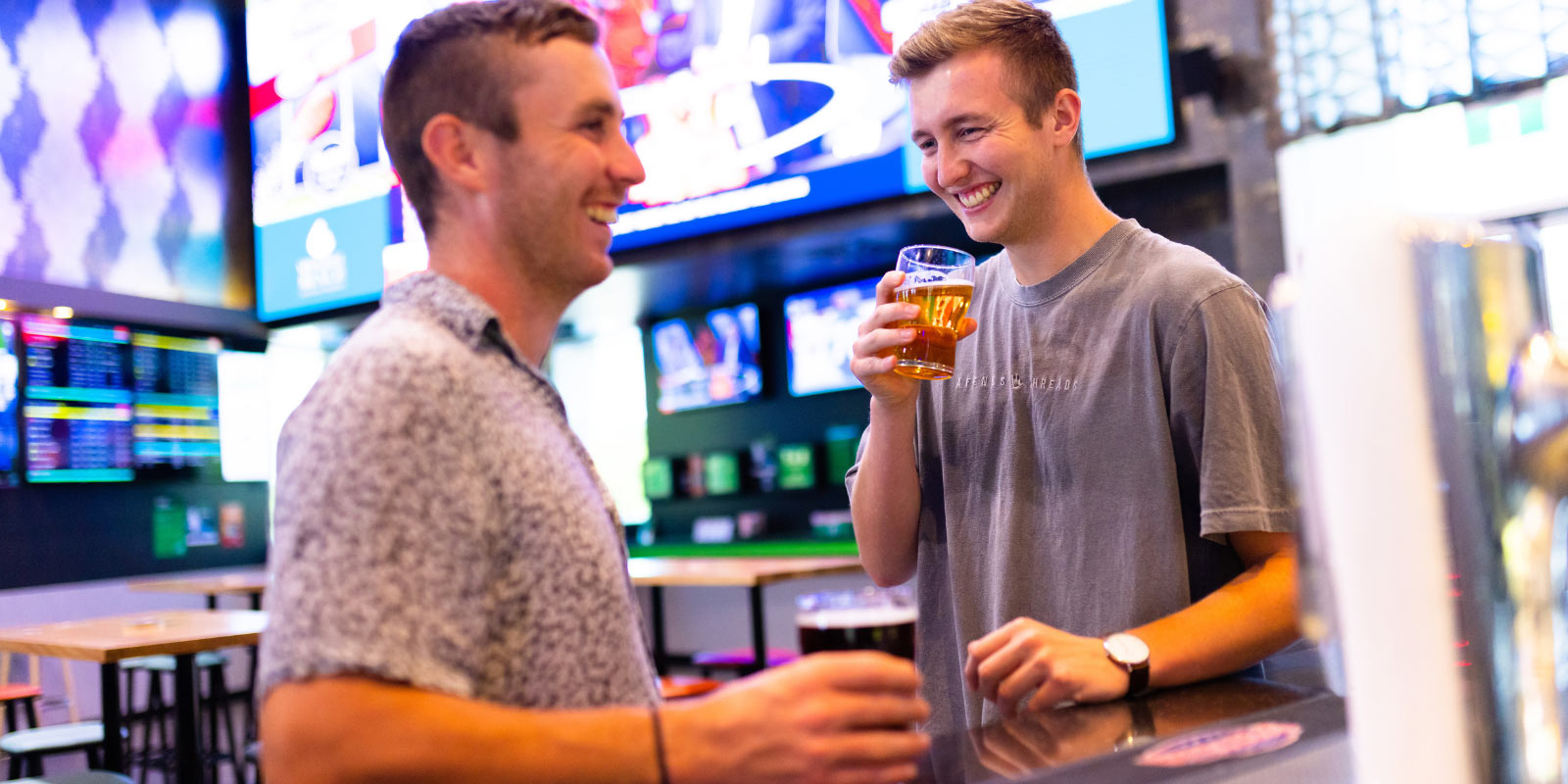 Two people at the bar counter with TV screens and digital signs behind them.